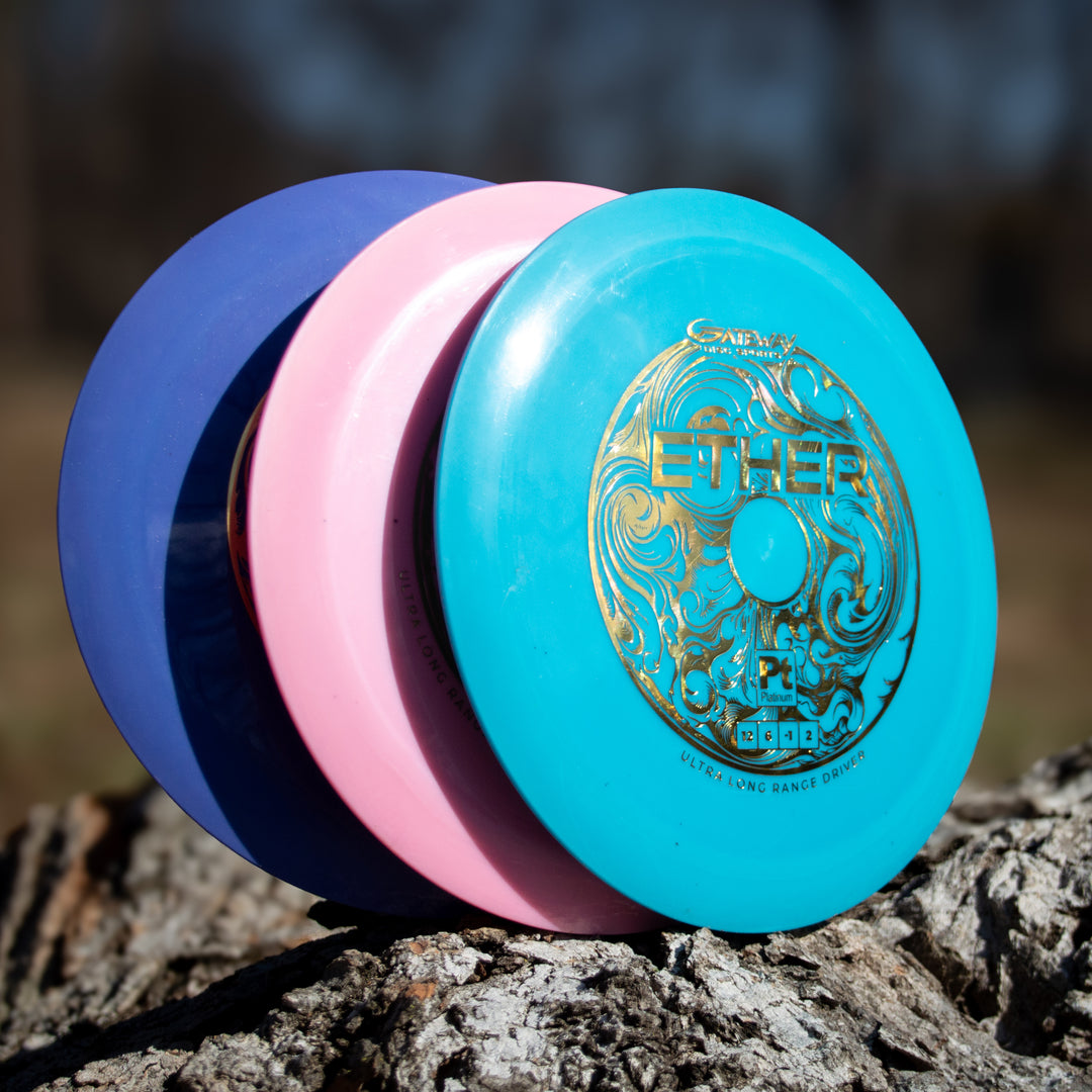 Three Ether - Platinum - New Run disc golf discs—blue, pink, and turquoise with gold design—are stacked upright on a tree stump outdoors, each made from eco-friendly plastic for superior grip and performance.