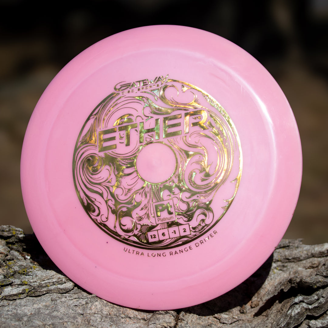 A pink Ether - Platinum - New Run distance driver with gold ornate lettering, made from eco-friendly plastic, rests on a piece of bark.
