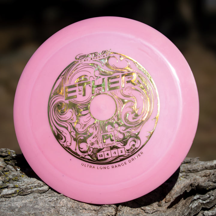 A pink Ether - Platinum - New Run distance driver with gold ornate lettering, made from eco-friendly plastic, rests on a piece of bark.