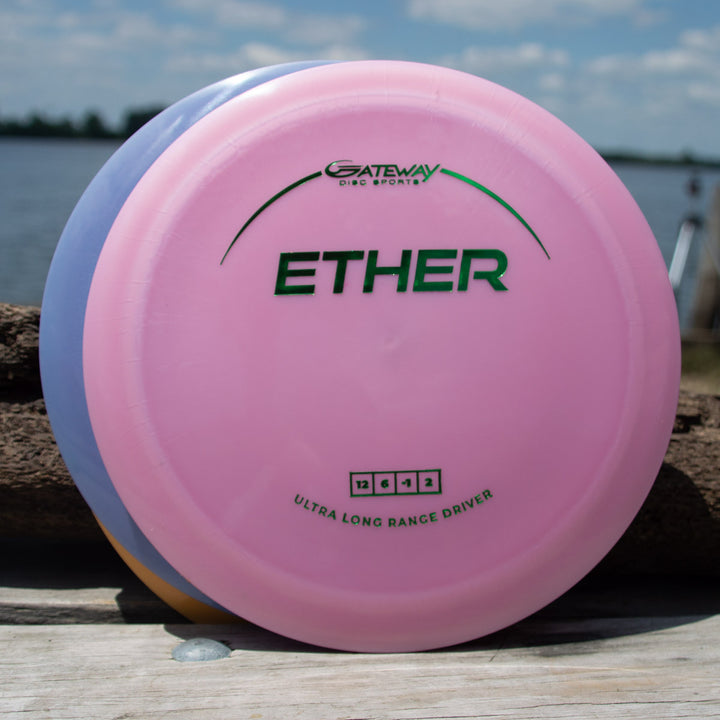 A pink Ether - Platinum - First Run distance driver by Gateway, made from eco-friendly platinum plastic, sits on a wooden surface outdoors with another disc behind it and a lake in the background.