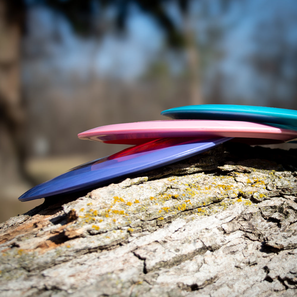 Three vibrant Ether - Platinum - New Run distance driver discs, crafted from eco-friendly plastic, stacked on a sunlit tree trunk with a blurred outdoor background.