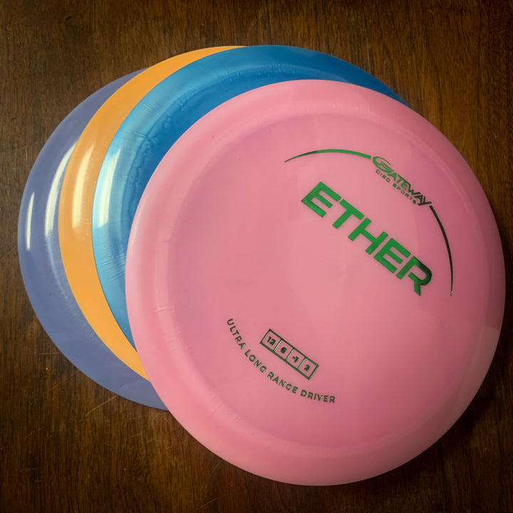 Three stacked disc golf discs in purple, blue, and pink rest on a wooden surface. On top is the Ether - Platinum - First Run distance driver, made from platinum plastic and featuring "ULTRA LONG RANGE DRIVER" on its label.