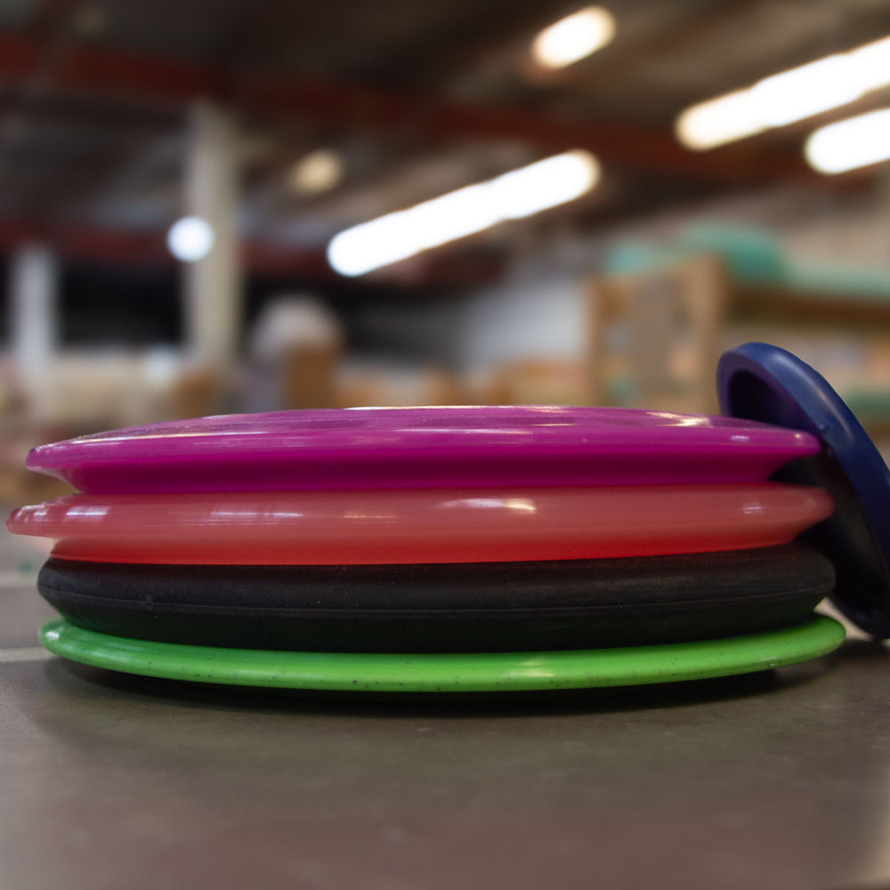 A Factory 2nd 4-Disc Pack, featuring colorful disc golf discs in pink, red, black, and green, sits on a table in an indoor setting.