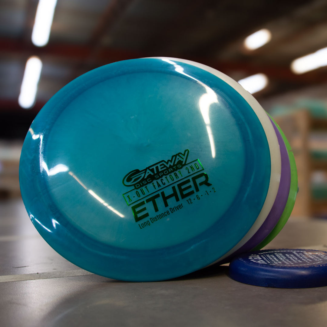 A blue Gateway ETHER distance driver from the Factory 2nd 4-Disc Pack stands upright on a table, with other discs from the set stacked nearby in a warehouse.