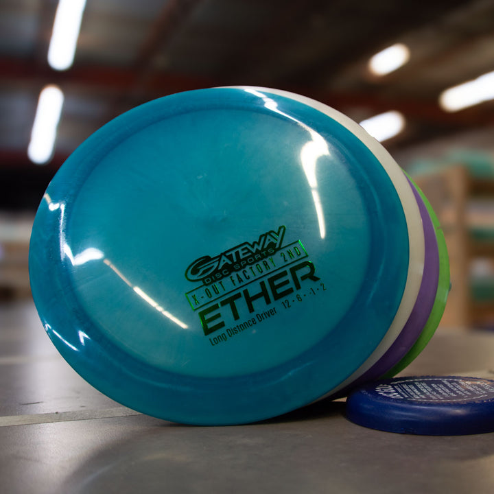A blue Gateway ETHER distance driver from the Factory 2nd 4-Disc Pack stands upright on a table, with other discs from the set stacked nearby in a warehouse.