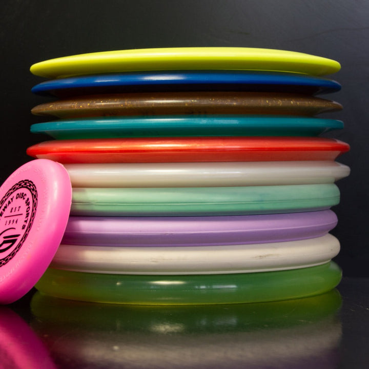 A stack of Gateway Factory 2nd Mystery 10-Pack discs, featuring vibrant colors and one pink disc propped upright, is displayed against a dark background.
