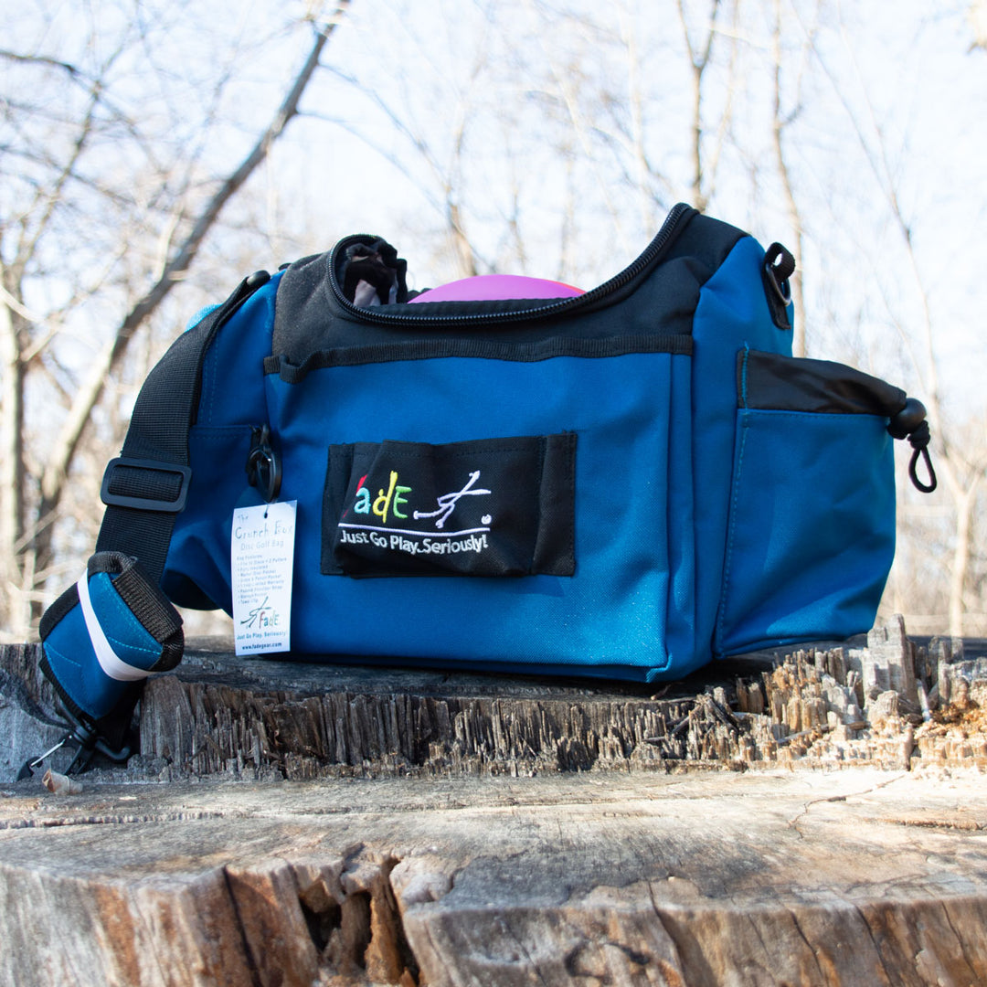 The blue Fade Gear Crunch Box Disc Golf Bag with a padded shoulder strap rests on a tree stump, featuring a "Fade Gear Just Go Play. Seriously!" patch—ideal for everyday disc golf play.