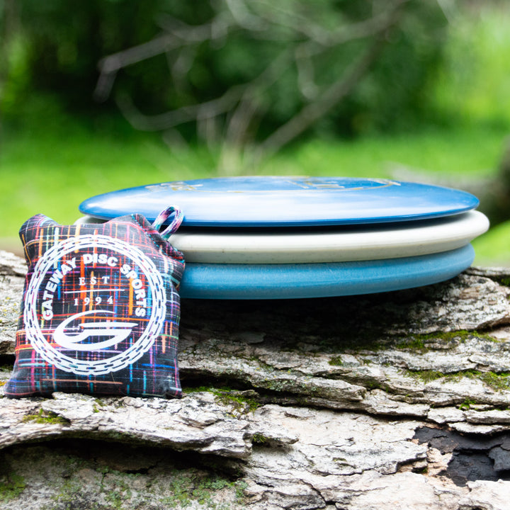 Three blue discs from the Father's Day Gift Set are stacked on a log outdoors next to a colorful bag with the "Gateway Disc Sports" logo. Green foliage is blurred in the background.