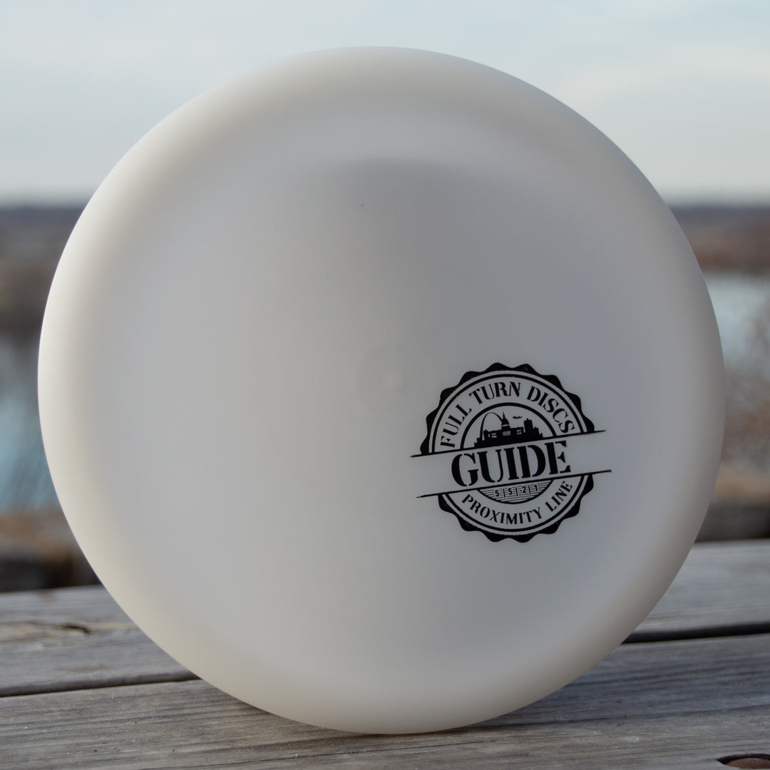 A white Full Turn Guide - Express disc golf disc with Flight Numbers and logo sits on a wooden surface outdoors.