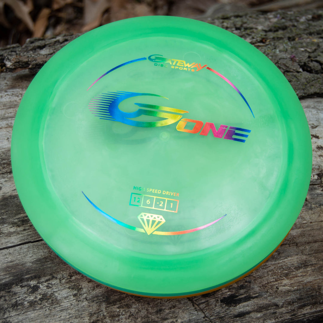 A green G-One distance driver from Gateway Disc Sports with rainbow lettering, designed for high speed turn, sits on a wooden surface outdoors.