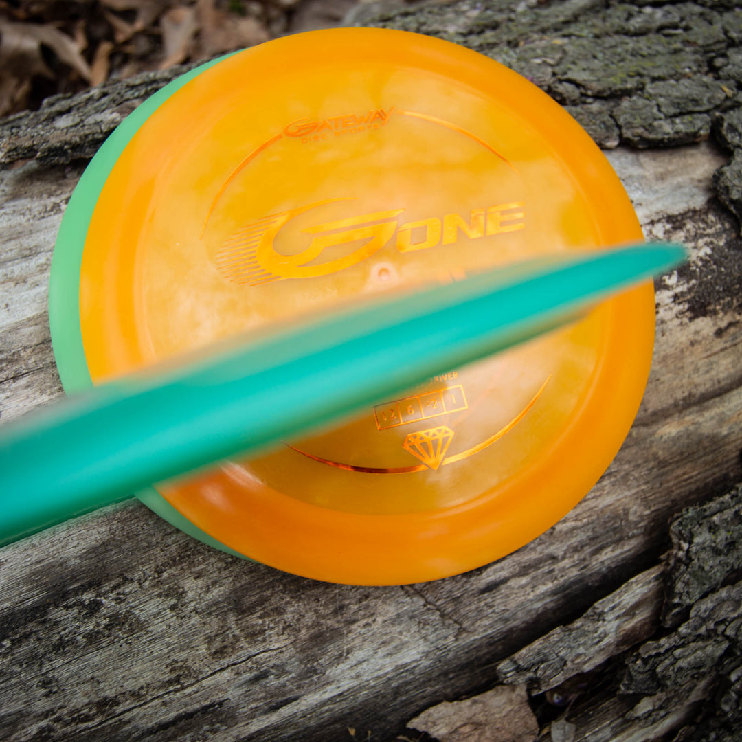 An orange G-One distance driver disc and a teal disc rest on a weathered log with textured bark.
