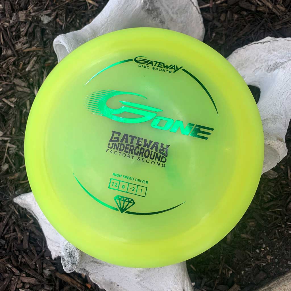A neon yellow G-One distance driver marked "Factory Second" sits atop white, bone-like structures with mulch in the background.