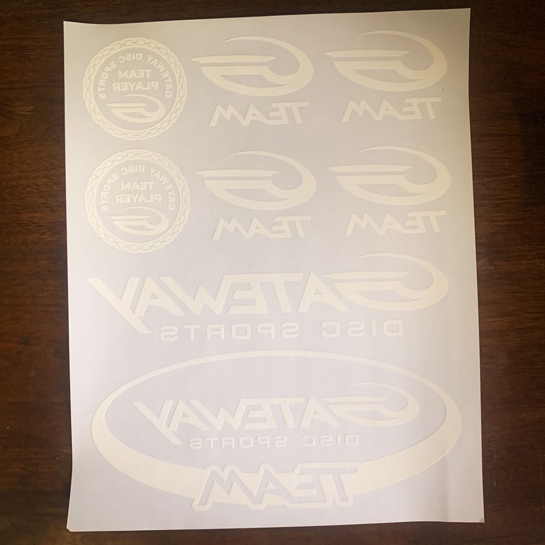 The Iron-On Team G Logo Sheet features white vinyl stickers with "Gateways Disc Sports Team Player" and "Team" logos—perfect for decorating gear or using with heat transfer paper. It is displayed on a dark wood surface.