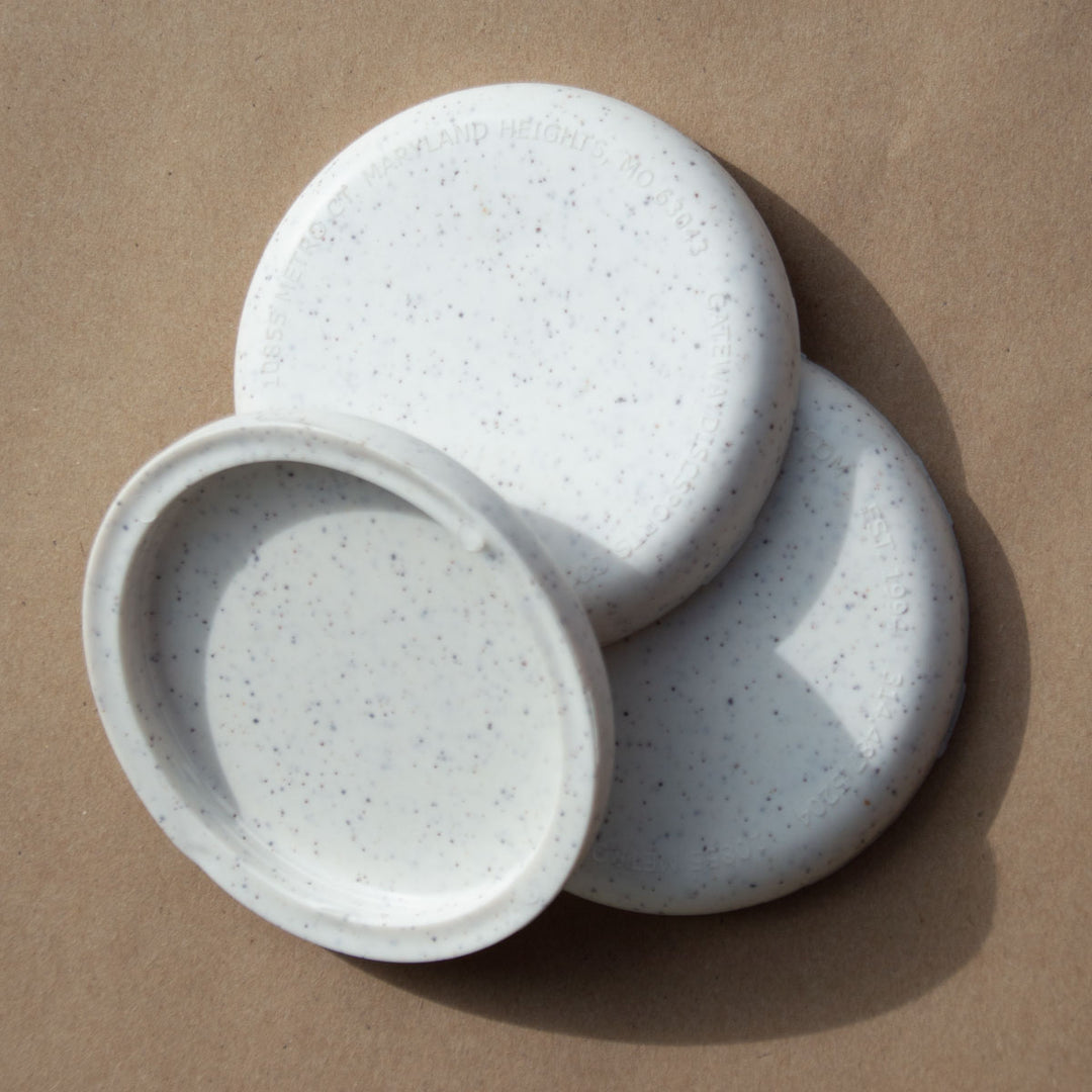 Three white, speckled, round Mini (Small) - Hemp - Blank lids are stacked on a brown paper surface. One is flipped to reveal the inside—ideal for custom stamp designs or as unique promotional giveaways.