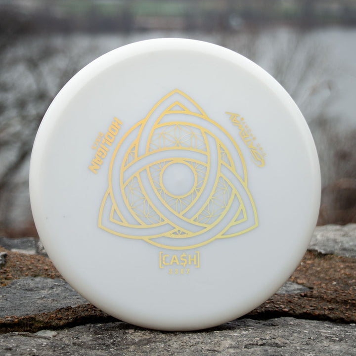 A white Hooligan CASH - Primo disc golf putter with gold geometric designs and "HOOLIGAN Discs," "Kinetic," and "[CASH] 2022" text sits on a rock, offering beaded precision and control for any player.