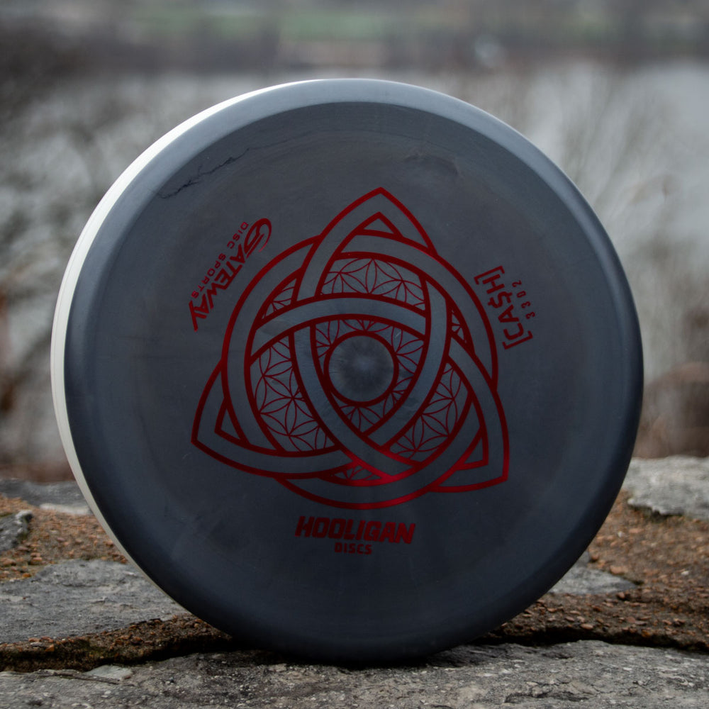 A gray Hooligan CASH - Primo beaded putter with a red geometric design and text stands upright on a stone surface outdoors, promising precision and control with blurred trees and water in the background.
