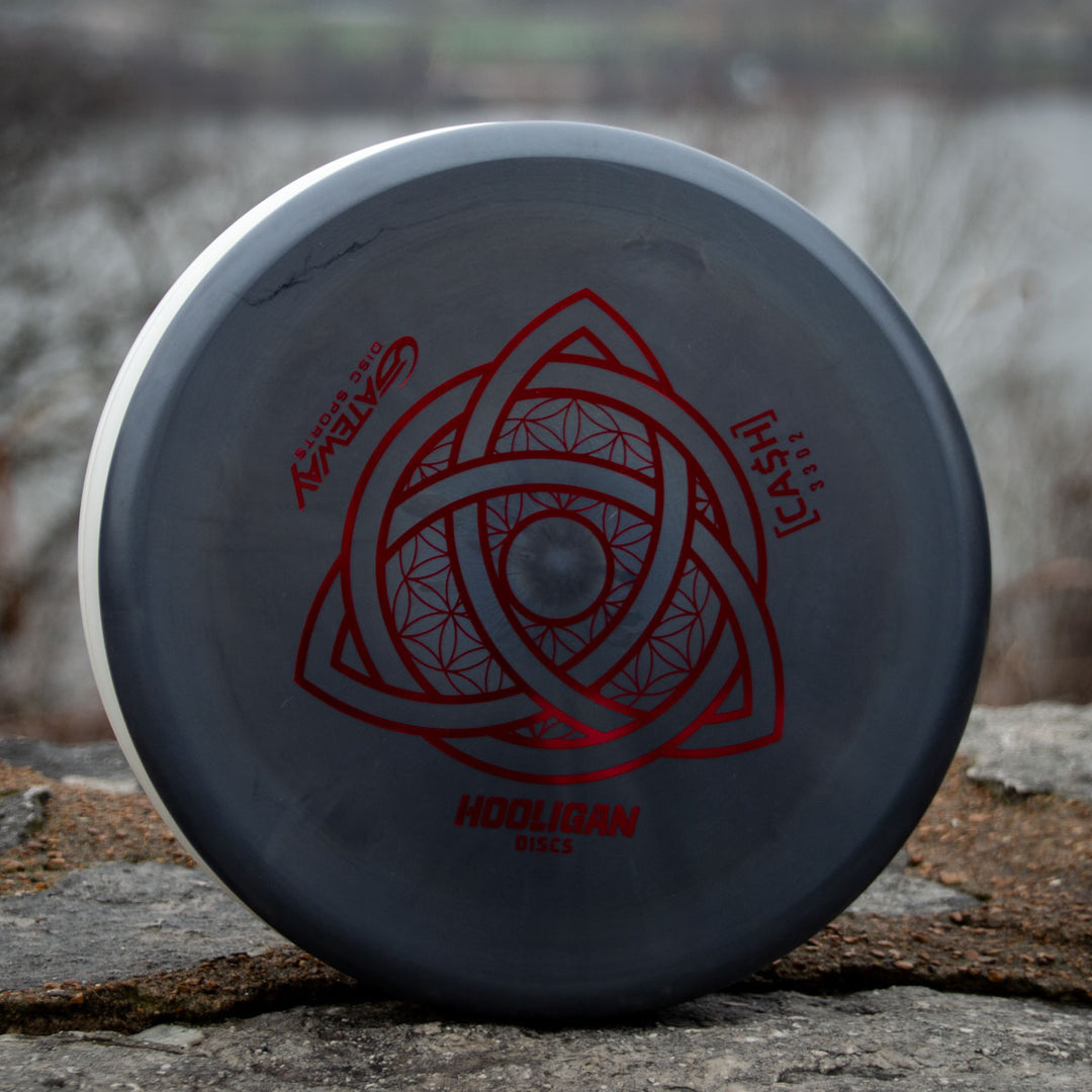 A gray Hooligan CASH - Primo beaded putter with a red geometric design and text stands upright on a stone surface outdoors, promising precision and control with blurred trees and water in the background.