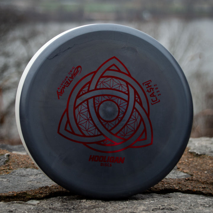 A gray Hooligan CASH - Primo beaded putter with a red geometric design and text stands upright on a stone surface outdoors, promising precision and control with blurred trees and water in the background.