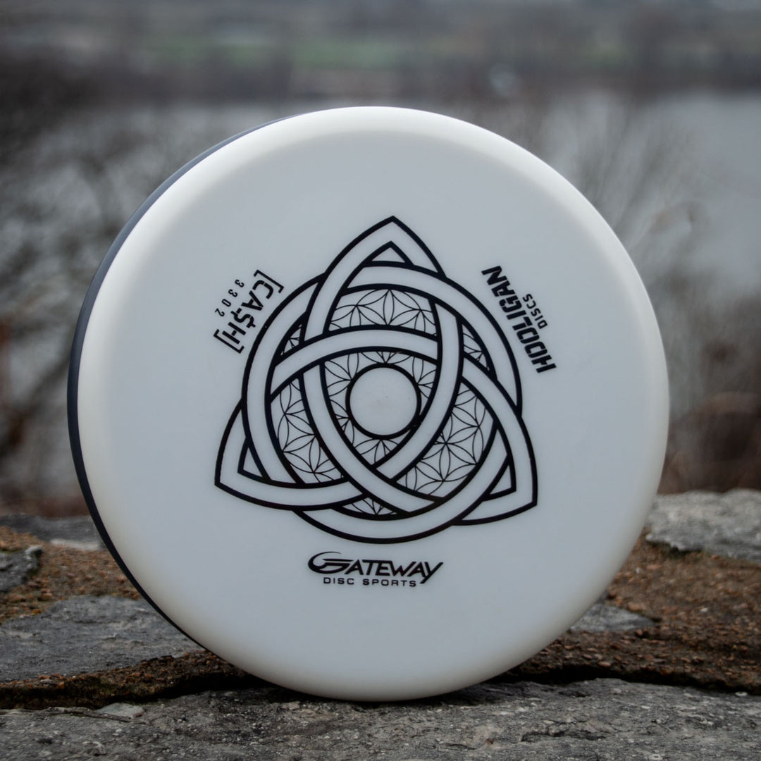 The Hooligan CASH - Primo is a white disc golf putter featuring a black geometric triquetra design and —CASH—� and —HOOLIGAN—� text, photographed outdoors on stone—a reliable, beaded option for players who value precision and control.