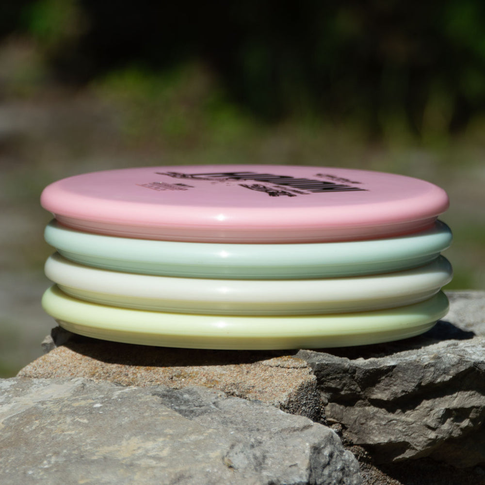 A stack of four pastel discs, including the Houdini - NXT Superglow putt and approach disc, sits on a rock outdoors, ready for power-gripping backhands.