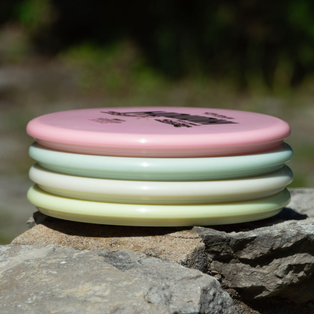 A stack of four pastel discs, including the Houdini - NXT Superglow putt and approach disc, sits on a rock outdoors, ready for power-gripping backhands.