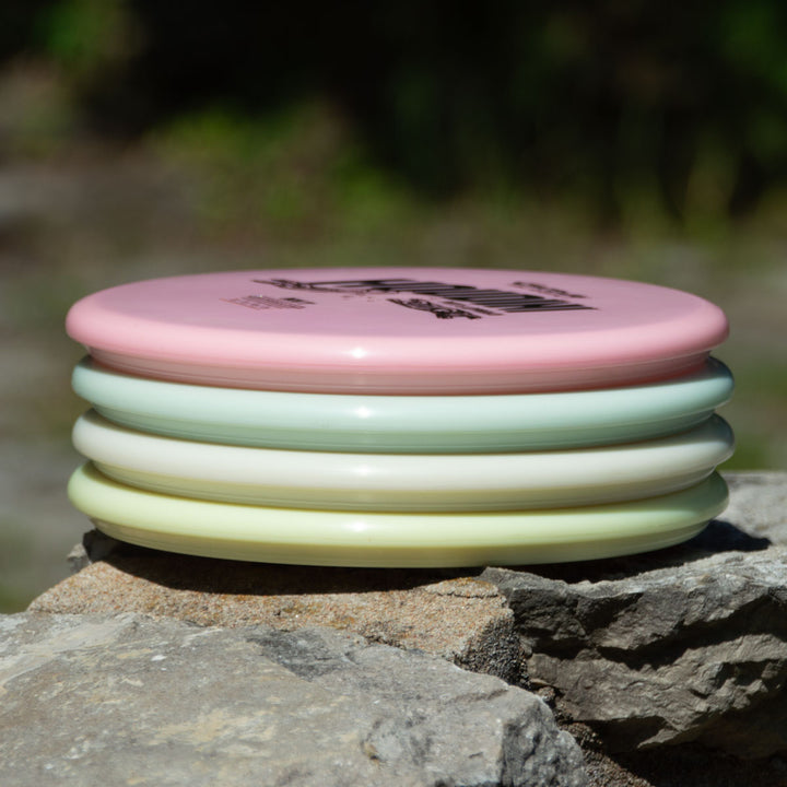 A stack of four pastel discs, including the Houdini - NXT Superglow putt and approach disc, sits on a rock outdoors, ready for power-gripping backhands.