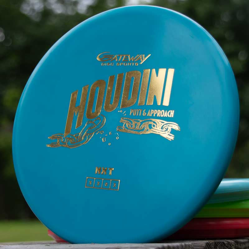 A blue Houdini - NXT putt and approach disc with gold "Gateway Disc Sports Houdini Putt & Approach" text stands upright outdoors, ideal for power-gripping backhands, with blurred trees in the background.