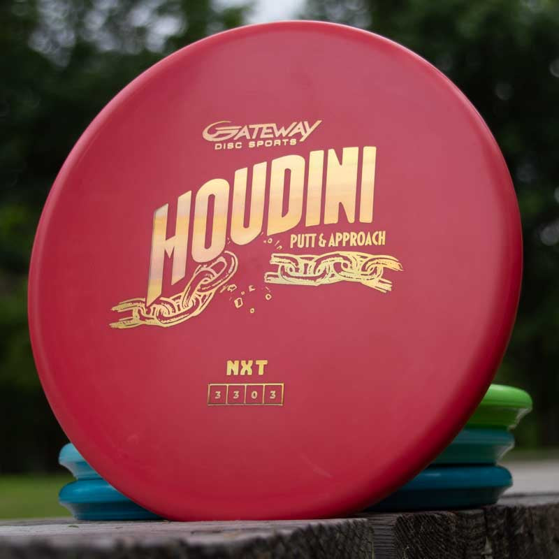 A red Houdini - NXT putt and approach disc by Gateway Disc Sports with gold lettering sits outdoors atop a stack of discs, ideal for power-gripping backhands.