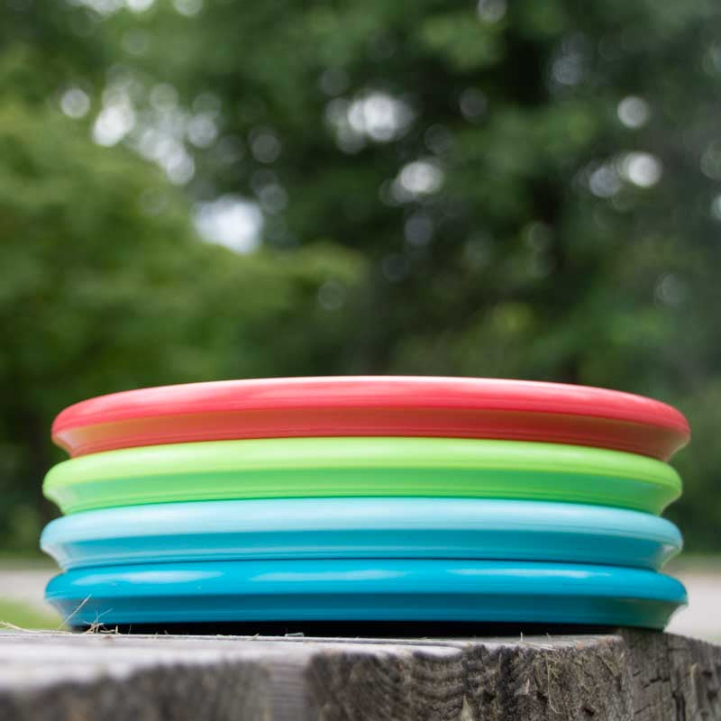 A stack of four disc golf discs—red, green, light blue, and blue, including the Houdini - NXT putt and approach disc—rests on a wooden surface outdoors with trees in the background for backhand practice.