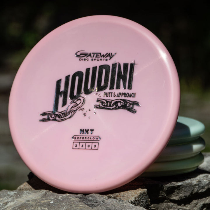 A pink Houdini - NXT Superglow disc from Gateway Disc Sports sits outdoors on other discs, ideal for power-grip backhands.