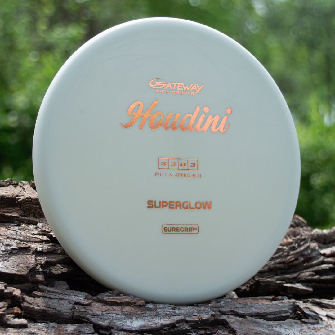 A white Houdini - SureGrip™ Superglow putt and approach disc with a deep rim leans against a tree trunk amid green foliage—ideal for power-grip backhands.