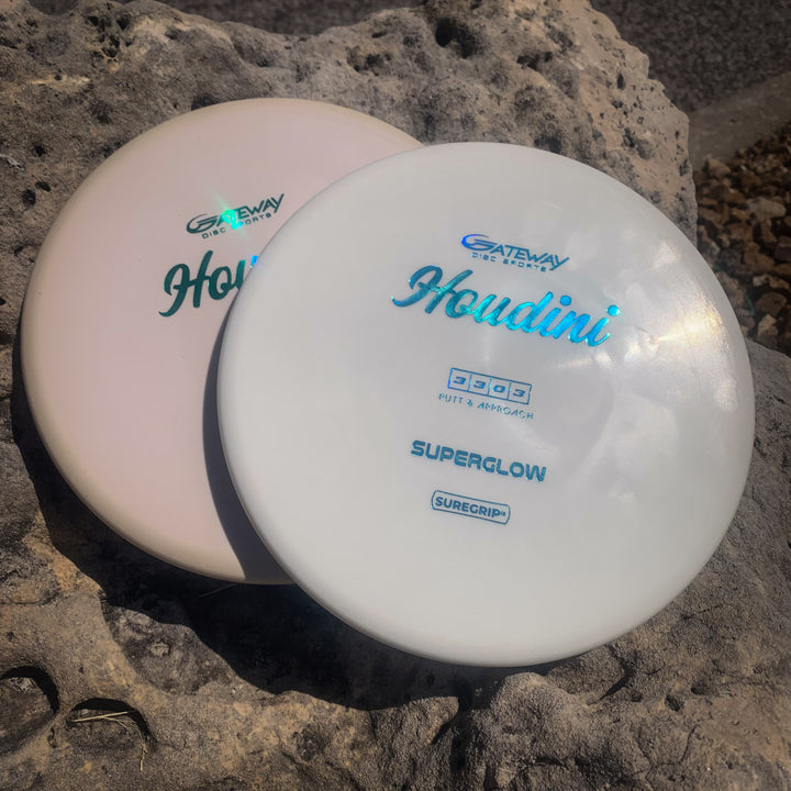 Two Houdini - SureGrip™Superglow discs, pink and white, with a deep rim, rest on a large outdoor rock. Designed for power-grip backhands or as putt and approach discs, each displays the “Superglow Suregrip” text.