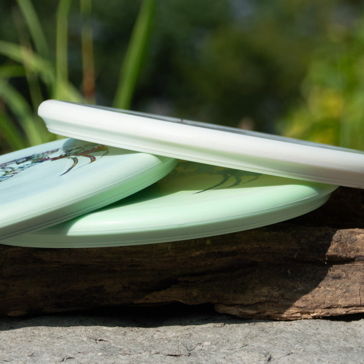 Three Brett Stetson Houdini - Superglow discs, featuring art by Kevin Hart, are stacked outdoors on wood with a blurred grassy background—ideal for your next disc golf tour.