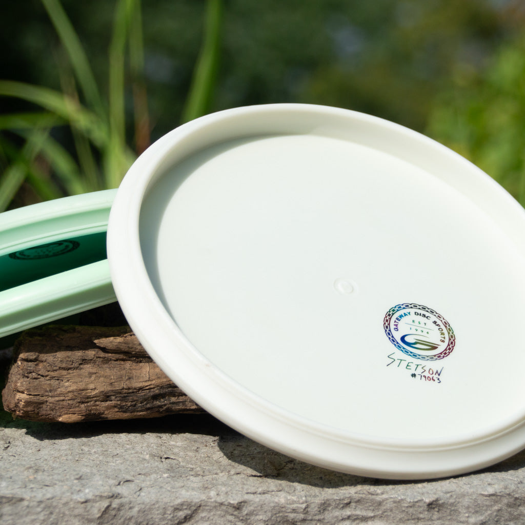 Two Brett Stetson Houdini - Superglow discs with Kevin Hart art rest outdoors on stone and wood, one featuring a central logo and text. Perfect for any disc golf tour.