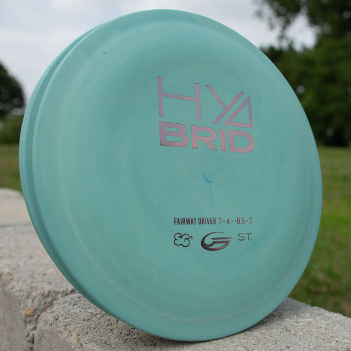 The Hybrid - Suregrip, a blue overstable disc golf disc, sits on an outdoor concrete surface with grass and trees in the background.