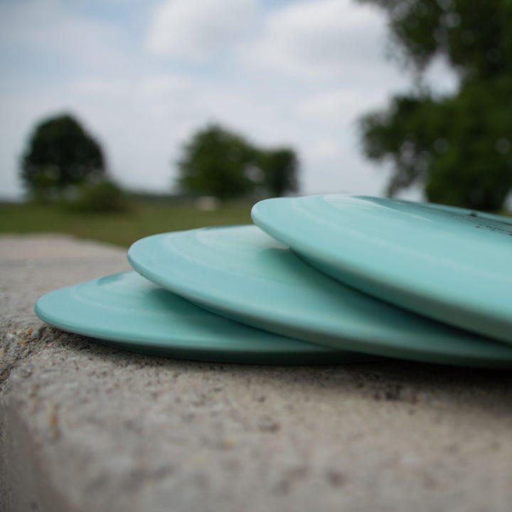 Three light blue Hybrid - Suregrip discs are stacked on outdoor concrete, with trees and a cloudy sky behind them. These discs feature advanced Surface Technology for overstable flight.