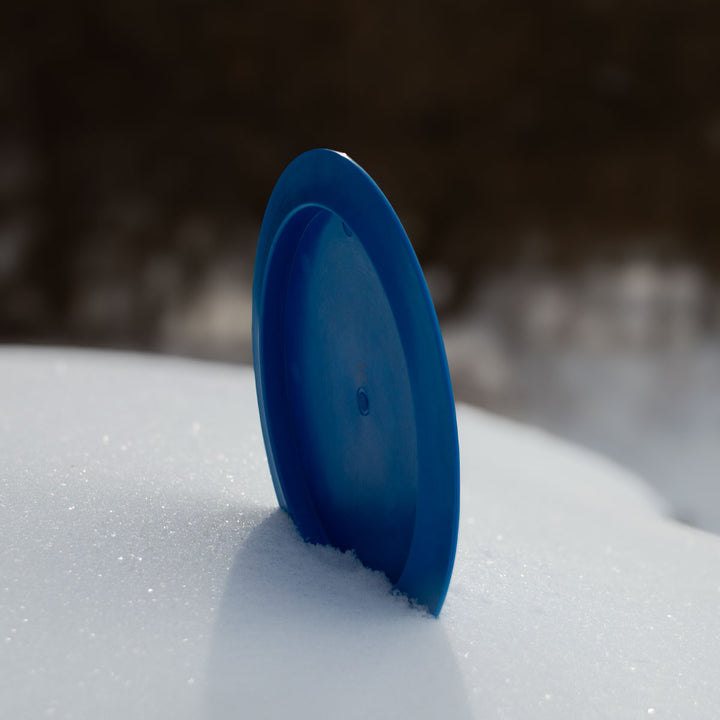 An Airstrike - Cobalt frisbee stands upright in white snow, its sleek disc silhouette set against a dark, blurred background.