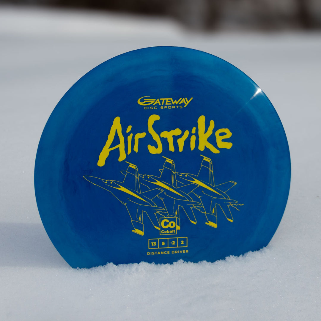 A blue Airstrike - Cobalt disc golf driver by Gateway Disc Sports, featuring yellow "Air Strike" text and three jet illustrations, stands upright on snow.