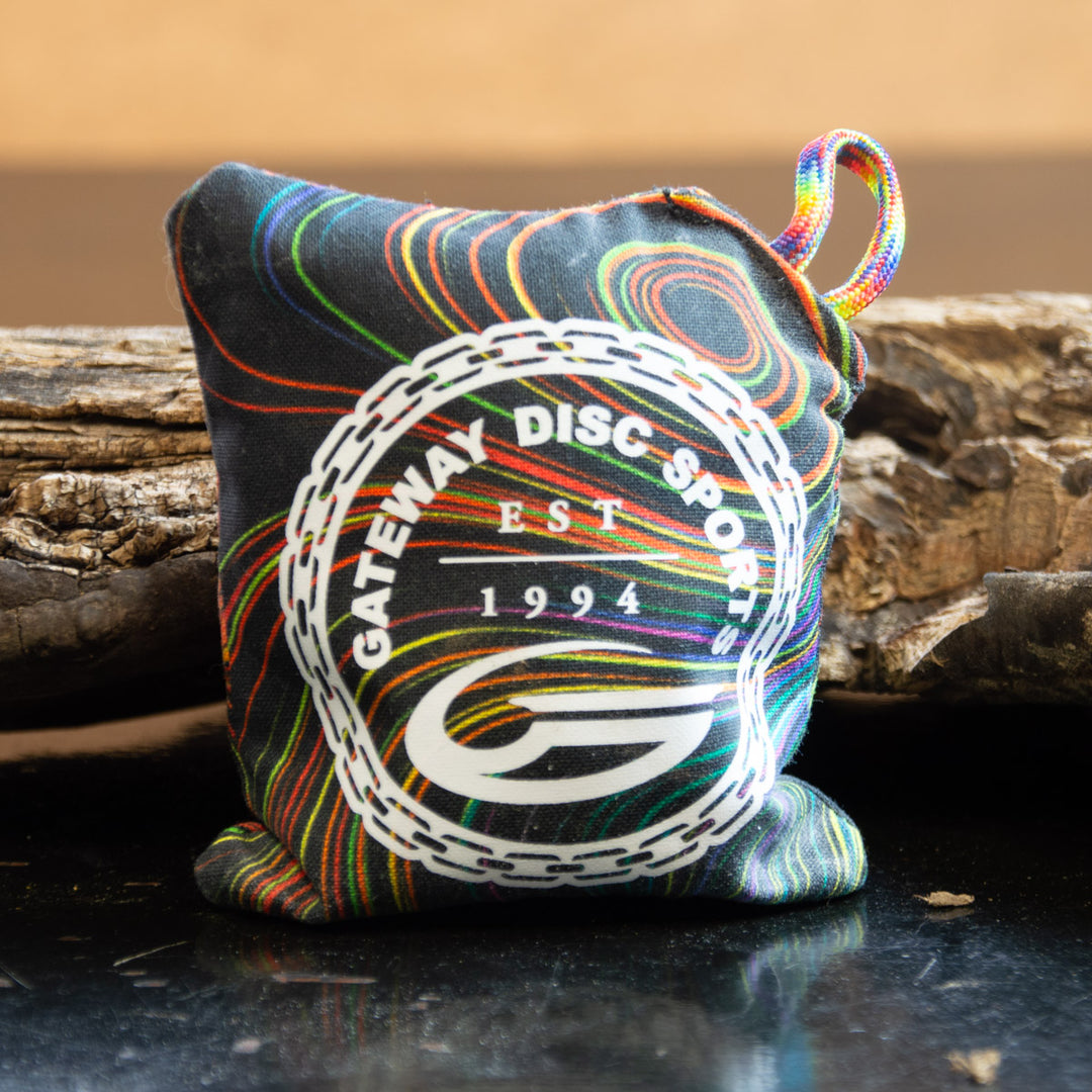 The Gateway Disc Sports x Jaybirdz Dry Bag €“ Grip-Enhancing Chalk Bag is a small, colorful pouch with the Gateway Disc Sports logo and "EST 1994" printed on it. It's designed for optimal chalk release and sits on a dark surface by a wooden piece.