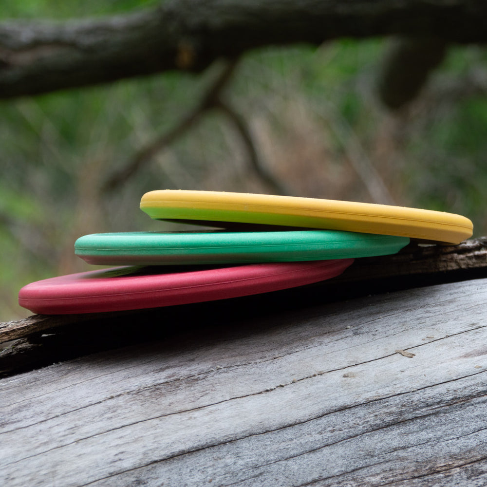 Three vibrant Jester DG - Wild Thing - Grippy Soft discs—yellow, green, and red—stacked on a weathered log with blurred trees behind; ideal for overstable approach shots or as a reliable beaded putt-and-approach disc.