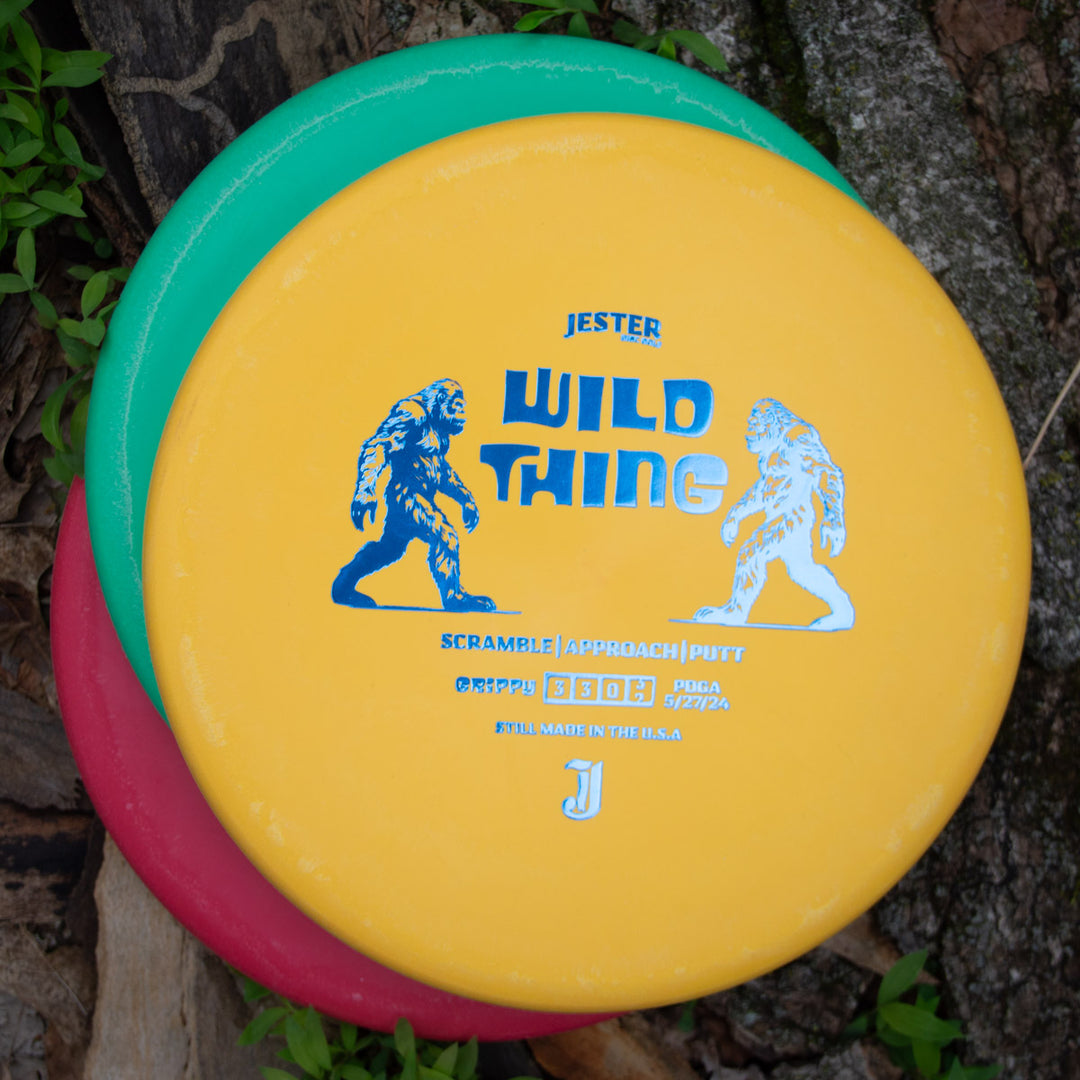 Three stacked Jester DG - Wild Thing - Grippy Soft discs, with the top yellow disc featuring Bigfoot art, rest on wood and leaves€”ideal as a beaded putt-and-approach disc for reliable overstable approach shots.