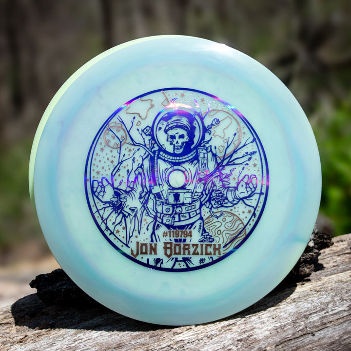 Jon Borzick's Ether - Color Superglow, a Gateway Disc Sports distance driver with a blue-ink skeleton astronaut and Jon Borzick's name on the front, made of NXT material, is displayed on wood outdoors.
