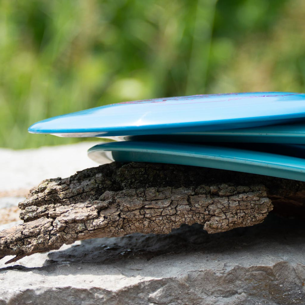 A blue Journey - Platinum distance driver disc golf disc rests on a piece of bark atop a stone surface with a blurred green background.