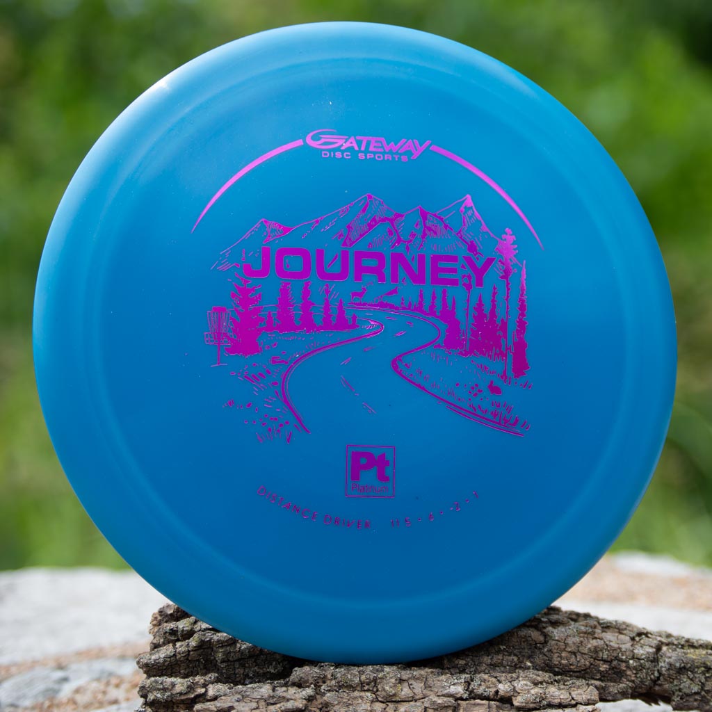 A Gateway Journey - Platinum disc golf disc, blue with a pink mountain and road graphic, sits on outdoor tree bark—this long-range distance driver is prized for its controllable flight.