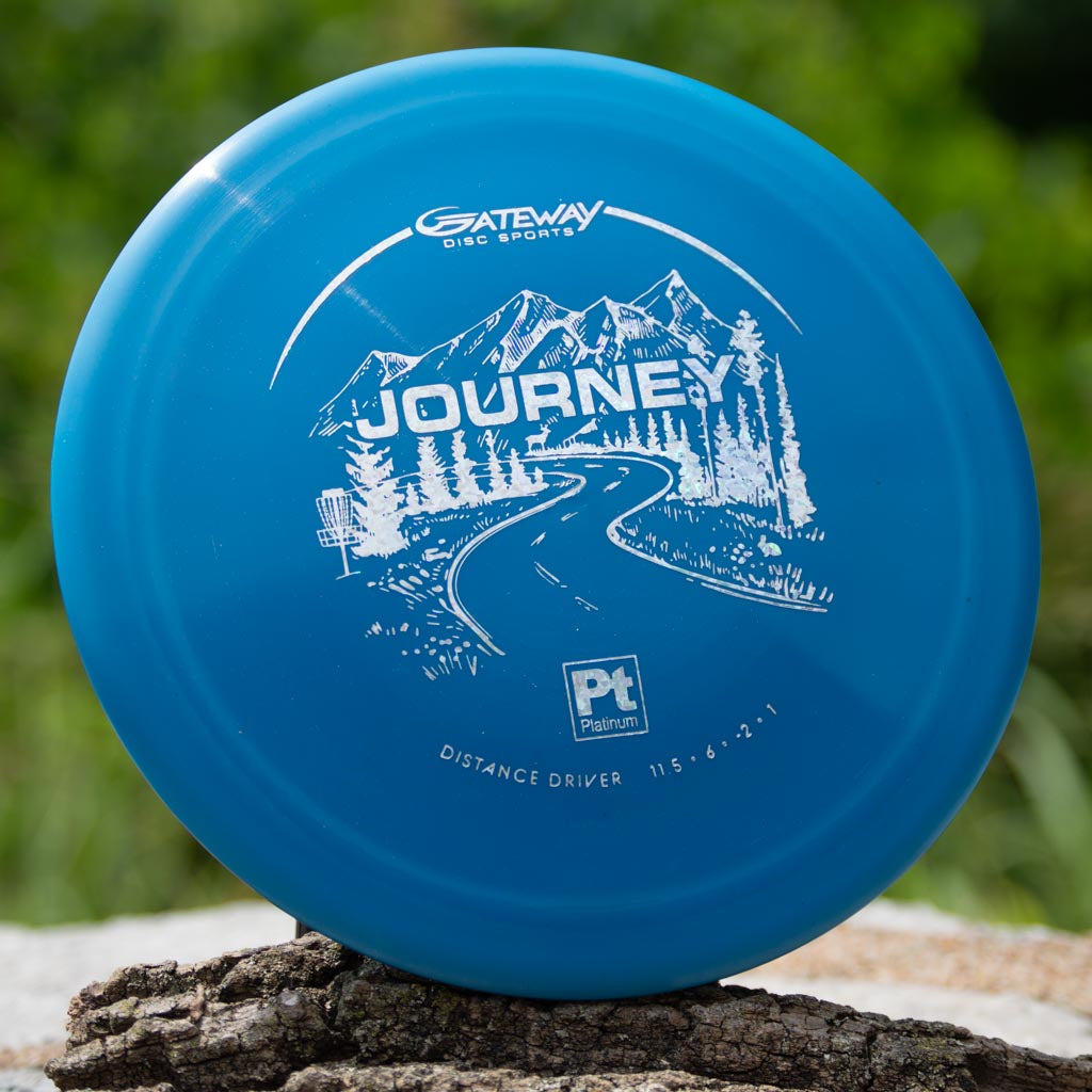A blue Gateway Disc Sports Journey - Platinum long-range driver with a white mountain, tree, and road graphic sits on wood outdoors—ideal for players who want controllable flight.