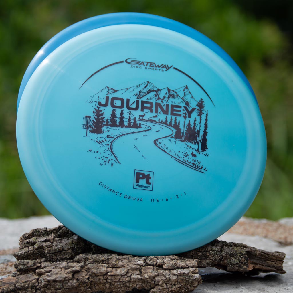 A blue Gateway Disc Sports Journey - Platinum long-range driver featuring a mountain and road graphic rests on bark outdoors, offering controllable flight and precision for players.