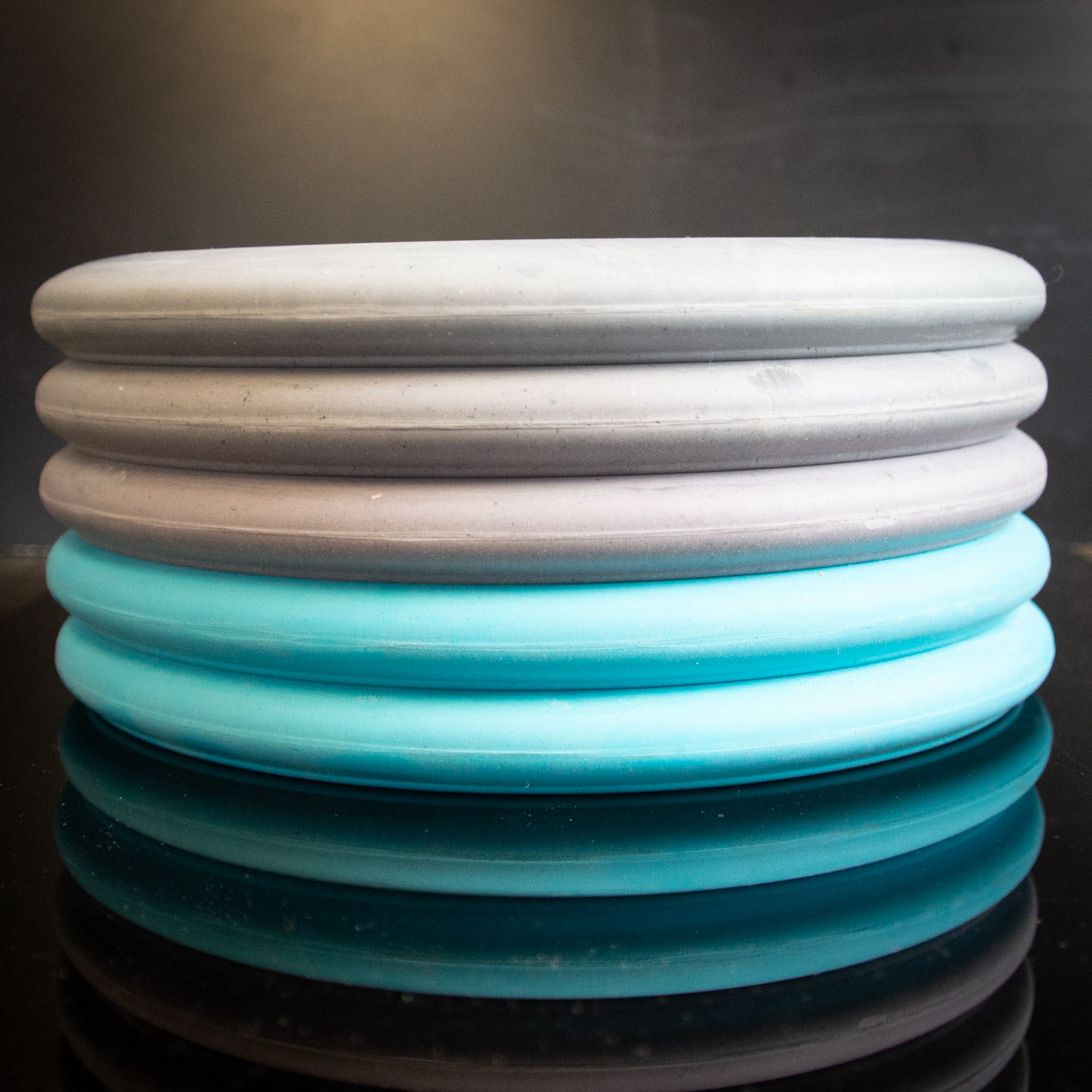 Four stacked circular disks in grey, purple, and blue shades with a ribbed texture—part of the Wholesale Monthly Promo Box disc selections—are displayed on a reflective dark background.