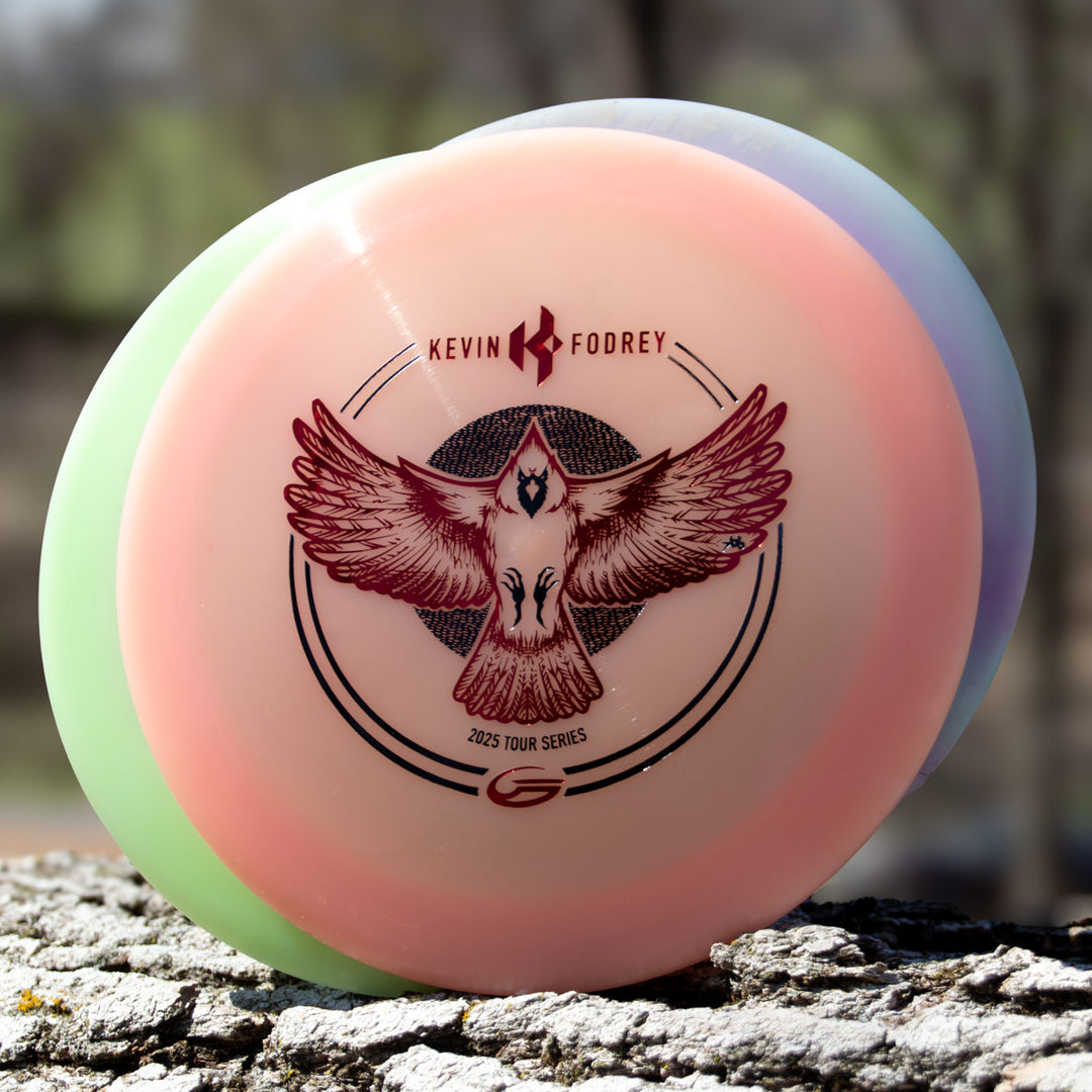 A pastel-hued Kevin Fodrey's Spell - Superglow overstable driver with a bird graphic and —Kevin Fodrey's Signature Spell 2023 Tour Series—� text rests outdoors on a textured surface.
