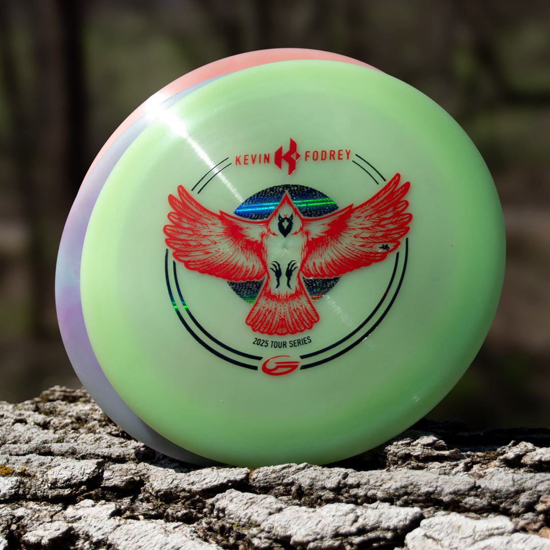 The Kevin Fodrey's Spell - Superglow, an overstable disc golf driver with a red bird graphic and "Kevin Foorley 2023 Tour Series" text, sits on a tree trunk outdoors, showcasing vibrant NXT Color Glow.