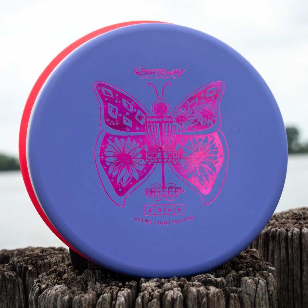 Lizzy's Chief OS - Ultra Lightweight, a blue and red overstable disc golf putter with a pink butterfly and basket design, stands upright on a weathered wooden post by the water—ideal for precise hyzer or forehand drives.